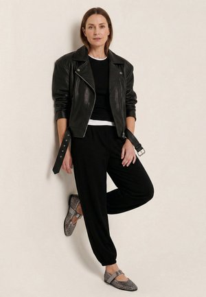 Vrouw met een zwarte leren jas, zwarte broek, zwart-witte gelaagde overhemden en grijze patroon Mary Jane platte schoenen, leunend tegen een muur.