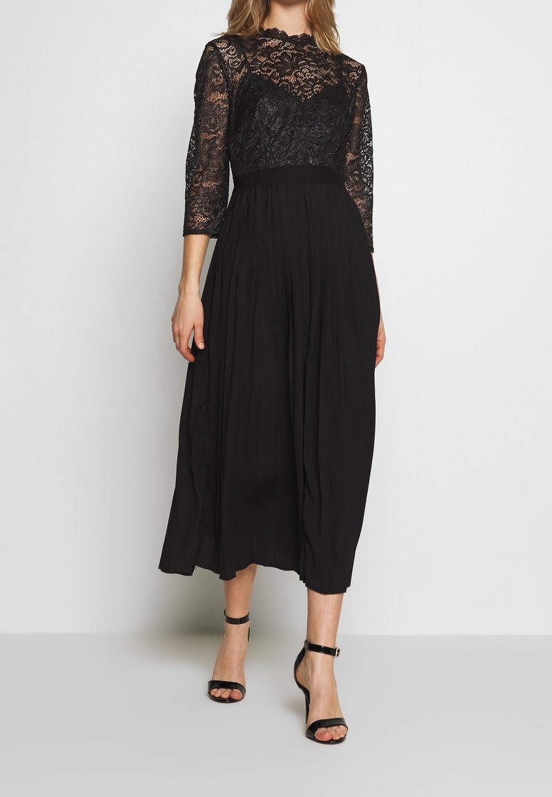 Femme portant une longue robe noire avec un haut en dentelle et des manches trois-quarts, associée à des sandales noires à talons hauts avec bride à la cheville.