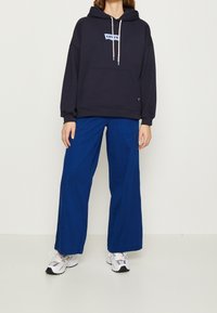 Tmavě modrá mikina s kapucí a modrým logem Levi's, s nastavitelnou šňůrkou u kapuce a přední kapsou, doplněná širokými elektricky modrými kalhotami a šedými teniskami.