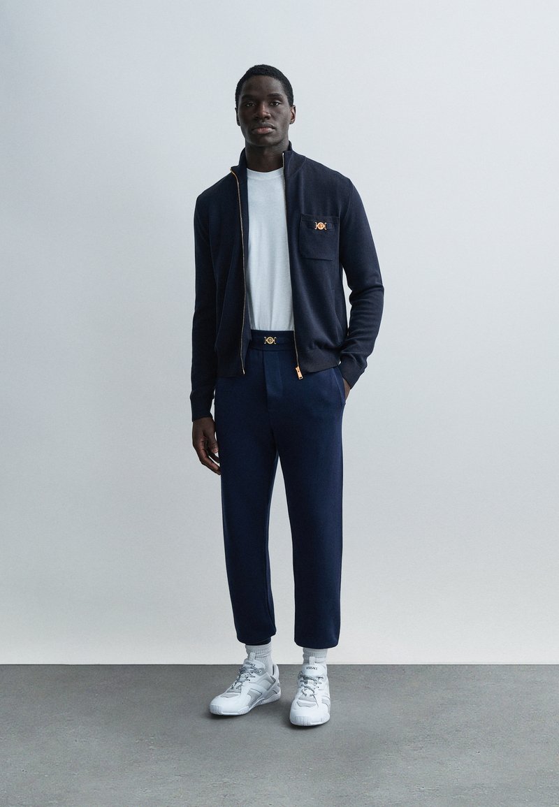 Homme noir debout, portant une veste zippée bleu marine et un pantalon assorti, une chemise blanche, des baskets blanches Versace et des chaussettes blanches, sur un fond clair uni.