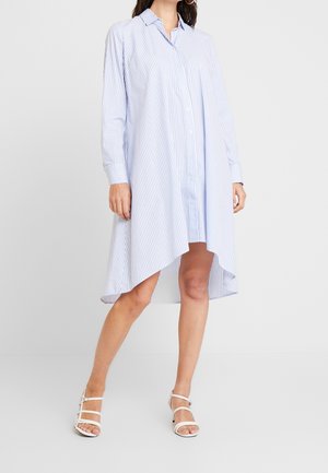 Frau trägt ein blau-weiß gestreiftes Hemdkleid mit langen Ärmeln und asymmetrischem Saum sowie weiße Riemchen-Sandaletten mit hohen Absätzen.