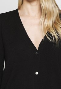 Czarny kardigan z dekoltem w serek, z dwoma guzikami — jednym czarnym i jednym białym. Miękka, teksturowana tkanina widoczna na zbliżeniu.
