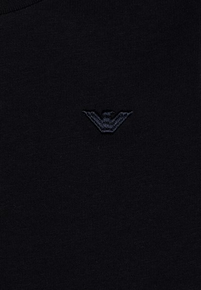T-shirt nera in maglia con scollatura tonda, dotata di un logo ricamato scuro sul lato sinistro del petto. Il tessuto ha una consistenza liscia.