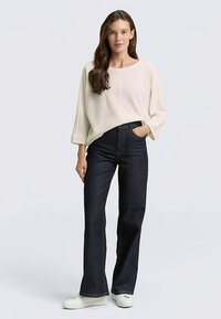 Maglione lavorato a maglia color crema chiaro con vestibilità ampia e maniche larghe, abbinato a jeans scuri a zampa di elefante a vita alta e sneakers bianche.