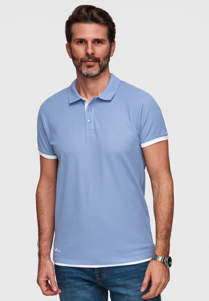 Homme aux cheveux courts et foncés, avec une barbe, portant un polo bleu clair à bordures blanches et un jean bleu, debout devant un fond uni.