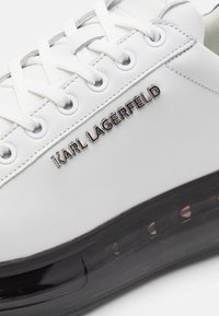 Balti odiniai sportiniai bateliai su apvaliu pirštu, turintys pilkus akcentus, su raišteliais priekyje ir juodu skaidriu padu, su įspaustu "KARL LAGERFELD" logotipu.