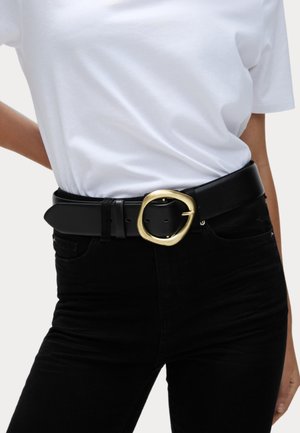 Cinturón de cuero negro con una gran hebilla ovalada dorada. El cinturón tiene una textura suave y se lleva sobre pantalones negros y una camiseta blanca.