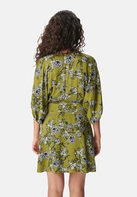 Vestido corto de tela verde oliva con un patrón floral en blanco y negro. Presenta mangas abullonadas y un detalle de botones en la parte trasera.