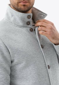 Chaqueta de algodón gris con un cuello alto, botones marrones y una textura suave. Camisa a rayas visible debajo.
