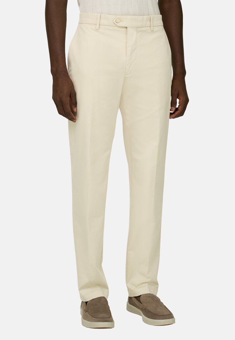 Homme portant un pantalon beige taille droite et des chaussures en daim marron à enfiler, debout devant un fond uni.