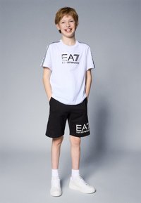 Camiseta blanca con logo negro "EA7 Emporio Armani", pantalones cortos negros con un logo similar y zapatillas blancas. Niño de pie con las manos en los bolsillos.