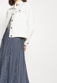 Bílá denimová bunda se dvěma náprsními kapsami, spárovaná s námořnickou sukni s kulatými bílými vzory. Outfit má uvolněnou siluetu.