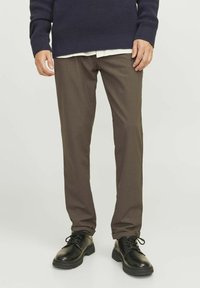 Braune Hosen aus einem glatten Stoff, kombiniert mit einem strukturierten, dunkelblauen Strickpullover und schwarzen Schnürschuhen. Saubere Linien und taillierte Passform.