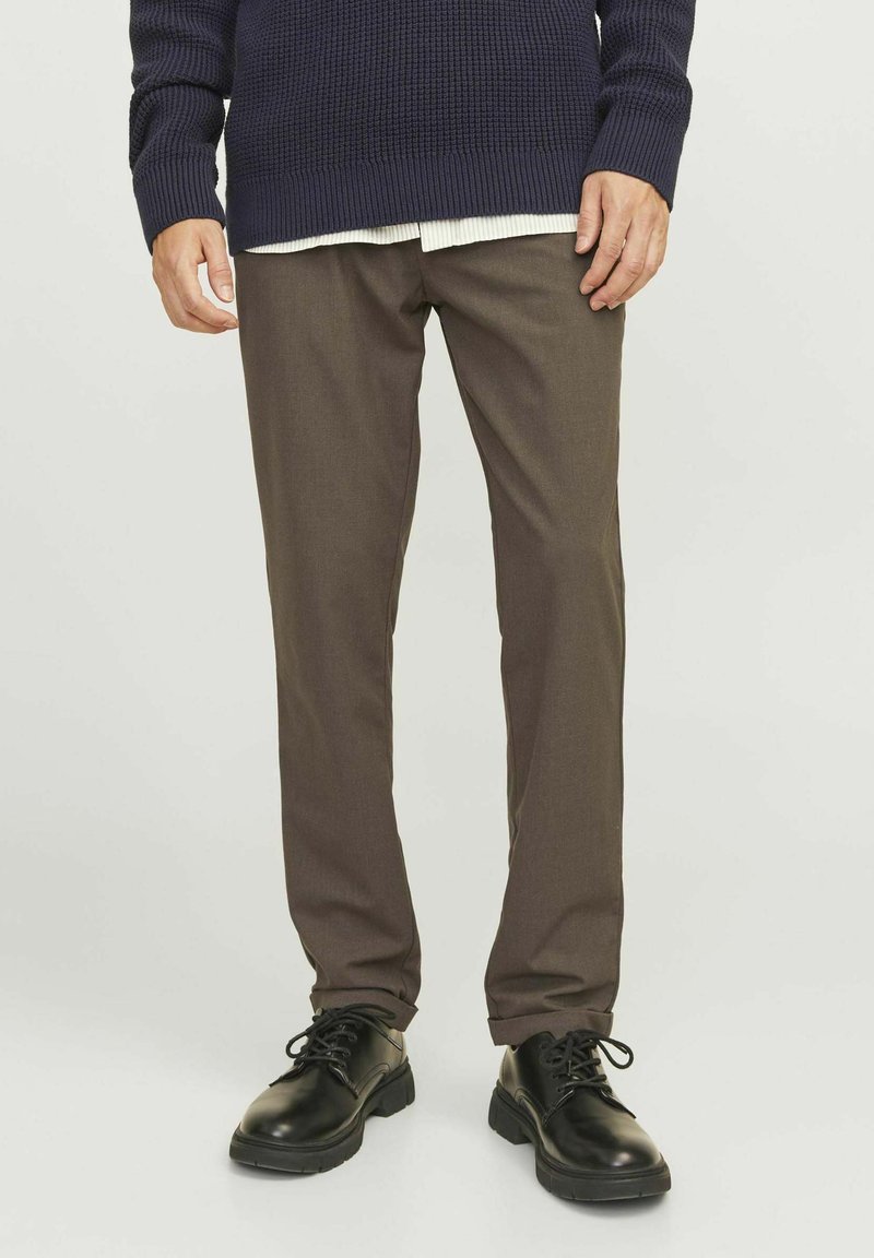 Braune Hosen aus einem glatten Stoff, kombiniert mit einem strukturierten, dunkelblauen Strickpullover und schwarzen Schnürschuhen. Saubere Linien und taillierte Passform.
