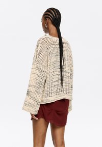 Gestrickter Pullover in Creme mit lockerem Schnitt, weiten Ärmeln und einem strukturierten Muster. Kombiniert mit einem kastanienbraunen Rock, von hinten getragen.