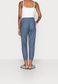 Pantalons jogger en denim avec une taille élastique, des chevilles fuselées et deux poches arrière, d'une couleur bleu moyen. Portés avec des sandales grises.
