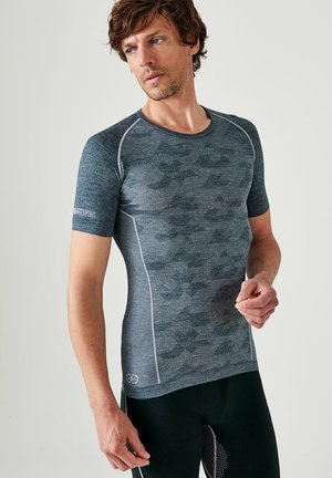 Man draagt een strak donkergrijs sportshirt met camouflagepatroon en zwarte leggings, kijkt naar links tegen een effen achtergrond.