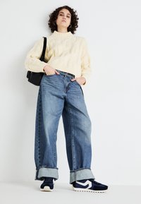 Pull en maille torsadée crème, jean en denim bleu loose avec des ourlets retroussés, et baskets navy avec des accents blancs, complété par un petit sac noir.