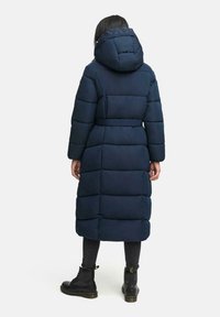 Lång, marinblå puffjacka med huva, bälte i midjan och stora quiltade sektioner. Bärs över mörka jeans och svarta snörstövlar.