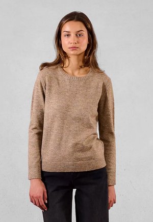 Donna con capelli castani lisci, che indossa un maglione di maglia beige e pantaloni neri, in piedi contro uno sfondo grigio chiaro uniforme.