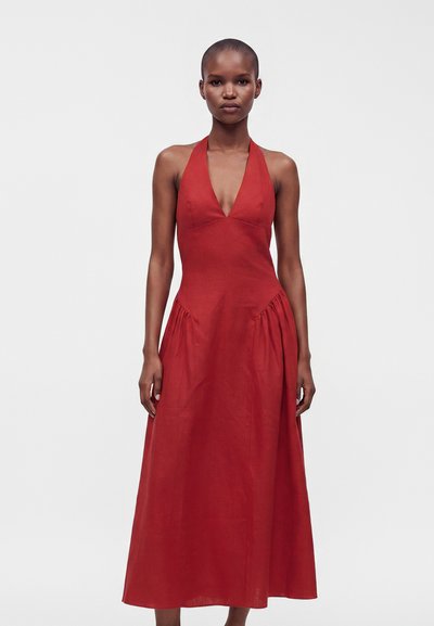 Woman wearing a sleeveless red halter-neck dress with a deep V neckline and a long, flowing skirt standing against a plain background.