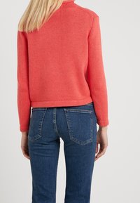 Pull tricot corail à col montant et poignets côtelés, associé à un jean en denim bleu foncé avec poches arrière et coupe ajustée.