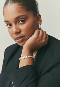 Silver braided bracelet worn on a wrist, featuring smooth, reflective surfaces and a twisted design. Black blazer with a notched collar.