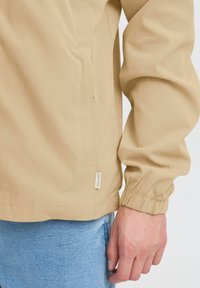 Beige lightweight jacket with elastic cuffs, side pocket, and a small brand tag. Smooth texture, designed for casual wear.