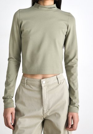 Kobieta w jasnym oliwkowo-zielonym krótkim golfie z logo "Calvin Klein" na kołnierzu oraz w beżowych spodniach z wysokim stanem zapinanych na guziki.