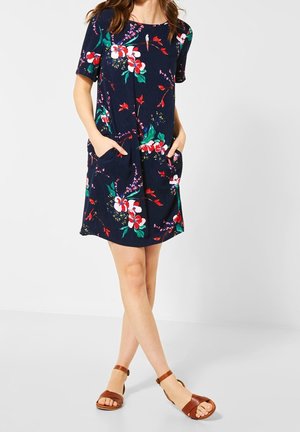 Femme portant une robe courte à manches courtes bleu marine à motifs floraux avec poches, debout les mains dans les poches, jambes croisées, et sandales marron à bride à la cheville.
