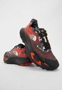 Chaussures de trail running avec un dessus en mesh rouge et noir orné de motifs, une semelle extérieure noire robuste et une marque bien visible sur le côté.