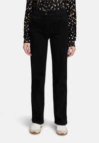 Persoon in zwarte rechte spijkerbroek, met een lang mouw leopardprint top en witte sneakers, staand met arme ontspannen langs het lichaam.