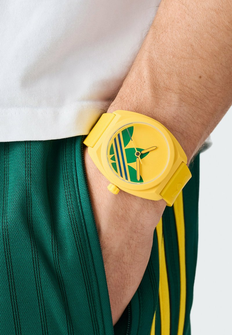 Gele plastic horloge met een ronde wijzerplaat, groene accenten en blauwe strepen; gedragen om een pols met een mouw van groen gestreepte stof.
