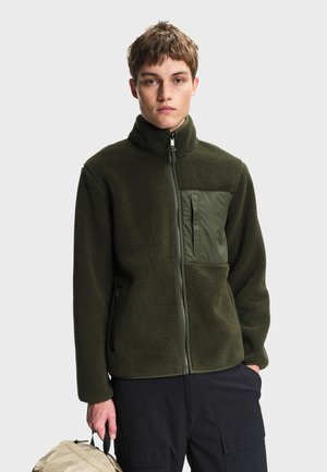 Jeune personne aux cheveux courts portant une veste polaire vert foncé, un pantalon noir, et tenant un sac à dos beige par la poignée.