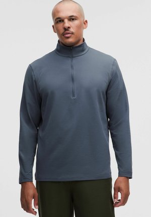 Man with shaved head wearing a long-sleeve dark gray quarter-zip pullover and dark green pants standing against a plain light background.