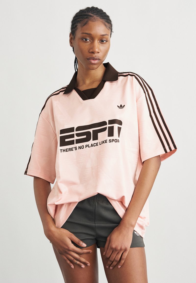 Femme aux cheveux tressés portant un grand t-shirt rose Adidas avec des rayures noires et un short noir, debout devant un fond uni.