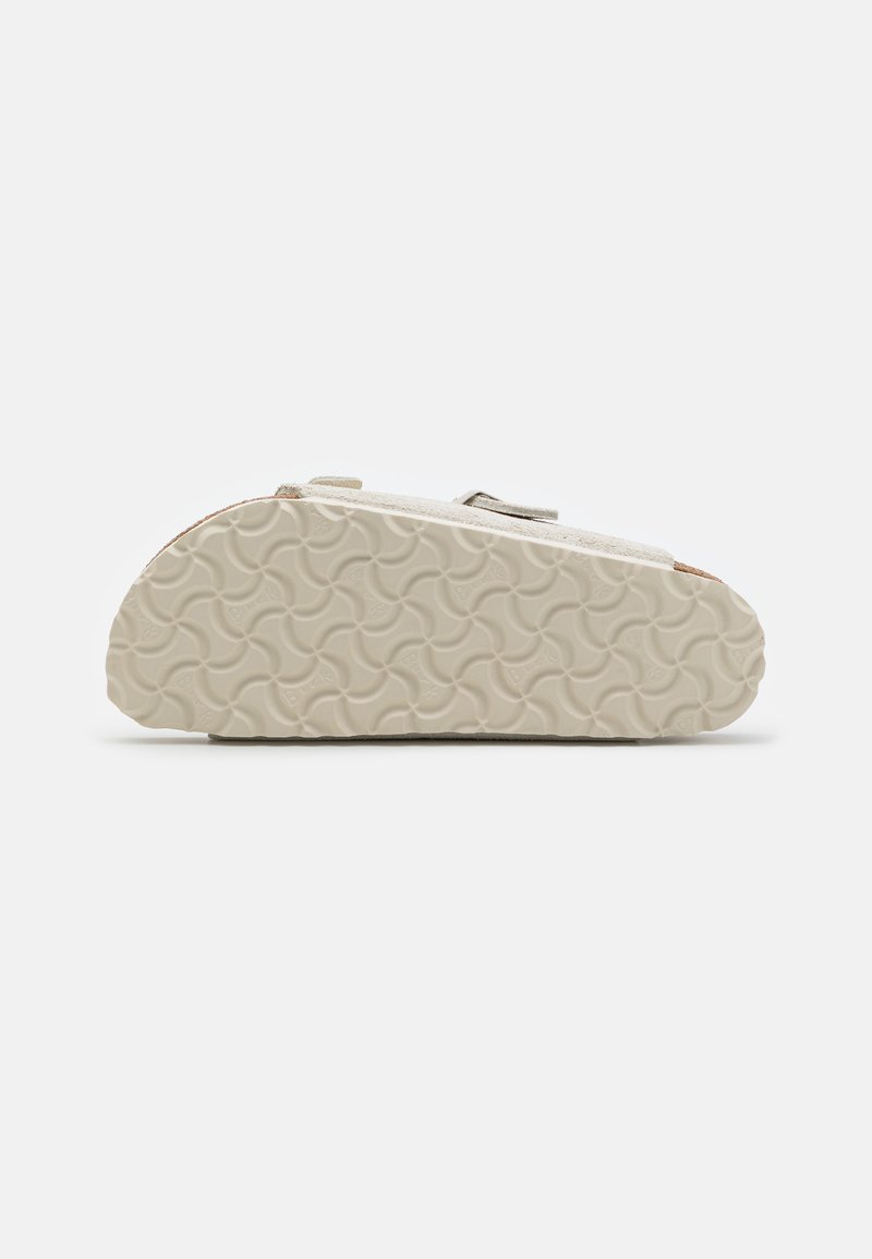 Helle graue Wildlederschuhe mit einer strukturierten, beigen Gummisohle, die ein wellenförmiges Muster aufweist, und zwei verstellbaren Klettverschlüssen für einen sicheren Sitz.