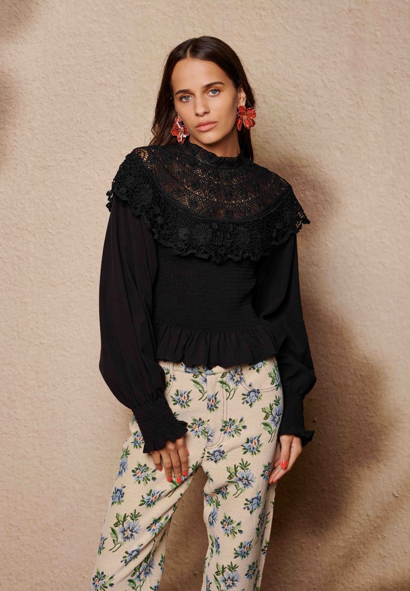 Blouse noire avec col à volants en dentelle et taille smockée, associée à un pantalon crème orné de broderies florales bleues.