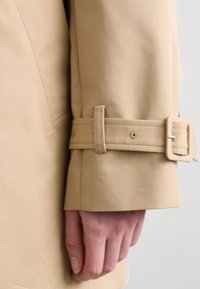 Main d'une personne portant un manteau beige avec une sangle ajustable et un détail de boucle sur la manche près du poignet.