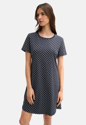 Kobieta w granatowej sukience z krótkim rękawem, ozdobionej małym okrągłym wzorem, stojąca na jasnym, jednolitym tle.