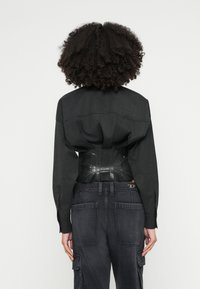 Camicia in tessuto nero con spalle strutturate, sovrapposta a un corsetto nero lucido. Abbinata a jeans scuri in stile cargo con tasche.