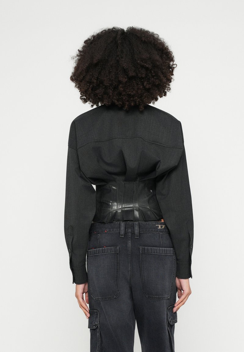 Schwarzes Hemd aus Stoff mit strukturierten Schultern, darüber ein glänzendes schwarzes Korsett. Kombiniert mit dunklen Cargo-Jeans mit Taschen.