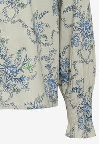 Blusa leggera a maniche lunghe di colore beige con motivi floreali blu e verdi, caratterizzata da polsini arricciati e una vestibilità rilassata.
