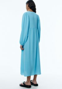 Blaues langärmliges Kleid aus strukturiertem Stoff, lockere Passform und geraffte Rückennaht. Kombiniert mit schwarzen flachen Sandalen.