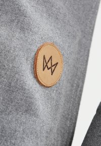 Tessuto grigio con una texture liscia, caratterizzato da un patch rotondo in pelle beige, impresso con un design di logo in marrone scuro.