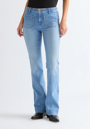 Vrouw draagt lichtblauwe bootcut jeans met voorzakken en zwarte schoenen met spitse neus, staand tegen een witte achtergrond.