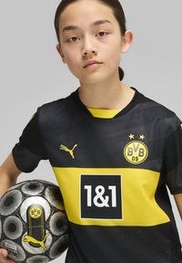 Camiseta negra y amarilla del Borussia Dortmund con el logo de Puma, de mangas cortas y con un patrón; sosteniendo un balón de fútbol negro y plateado.