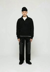 Black knitted pullover with a collar, paired with black trousers featuring glossy side panels. Model wears a black beanie and sneakers.