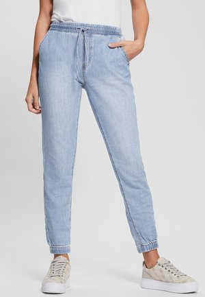 Osoba nosząca jasnoniebieskie spodnie dresowe typu jogger z elastycznym pasem i ściągaczami przy kostkach, w połączeniu z białymi, niskimi trampkami z wzorem.