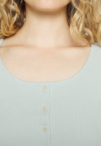 Světle modrý žebrovaný top má výstřih do tvaru lodi a tři odpovídající knoflíky pro zapínání vpředu, vyrobený z jemné texturované látky.